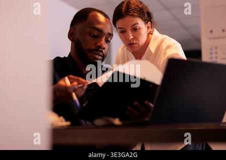 Project manager helping coworker checking report statistics, comparing data on laptop in office at night. Two employees checking marketing research results together in coworking space Stock Photo