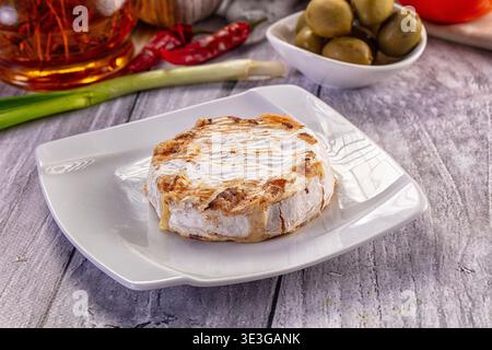 A wheel of golden-brown grilled Camembert cheese served on a modern white plate. The rustic scene includes olives, peppers, and green onion on a gray Stock Photo