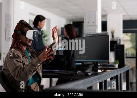 African american system engineer wearing virtual reality goggles working to develop security program in it big data office, software developer with vr headset coding html script with database. Stock Photo