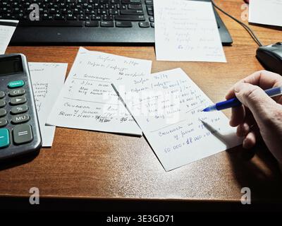 Kirov, Russia - February 02, 2026: A hand writes long-distance travel notes on cards on a wooden desk with a calculator and papers scattered around. Stock Photo
