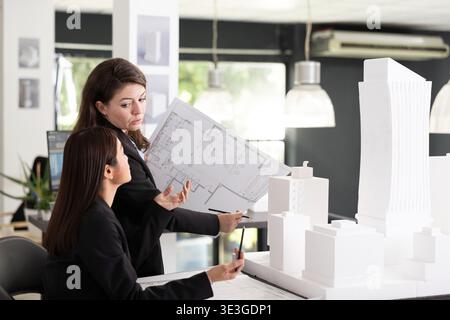 Caucasian manager explains project floor plans to coworker while examining white architectural model. Female design team collaborating in office, planning and ensuring successful construction project. Stock Photo