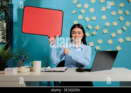 Professional asian woman with advertising speech bubble board against blue wall, showing isolated cardboard text message. Corporate employee holding poster with copy space. Stock Photo