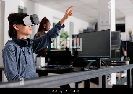 Asian software developer using virtual reality goggles to code new server interface, analyzing script information on terminal window. Coder programming it database for internet software with vr headset. Stock Photo
