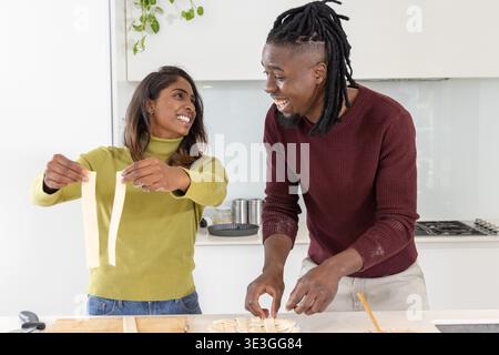 Diverse couple preparing fresh pasta at kitchen island with wooden board, mixing bowl Stock Photo