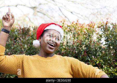 African American woman posing by trimmed hedge in Santa-hat and mustard sweater, pointing, smiling Stock Photo