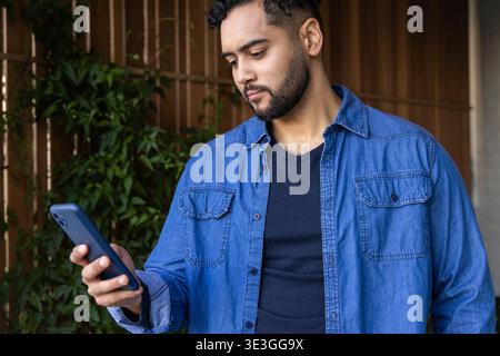 Man standing on shaded patio holding smartphone and wearing denim shirt near wooden fence Stock Photo