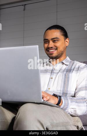 Asian man on sofa at home, using laptop, smiling, wearing striped shirt, khaki pants, watch, blinds Stock Photo