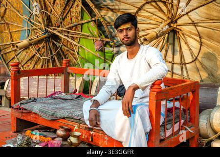 A young priest sits on a red wooden bench in front of large bamboo umbrellas at Dashashwamedh Ghat in Varanasi by the River Ganges. Stock Photo