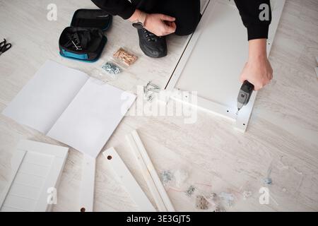 Top view of person hands using cordless drill to assemble white wooden furniture. Stock Photo