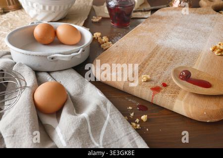 Baking preparation with brown eggs, whisk and wooden spoon on floured board. Close-up shot of rustic kitchen scene featuring fresh eggs in ceramic Stock Photo