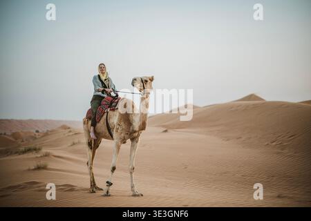 Caucasian woman wearing hijab rides camel in middle east desert Stock Photo