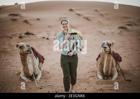 Woman in hijab laughs while walking between two camels Stock Photo