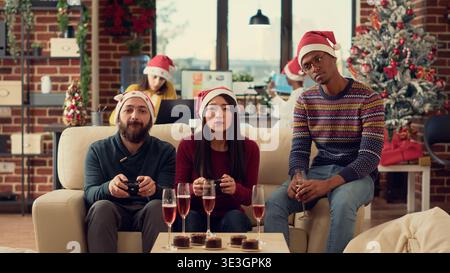 Diverse coworkers playing video games on console during christmas party celebration in office. Using controller to play gaming competition with colleagues, celebrating winter holiday. Tripod shot. Stock Photo
