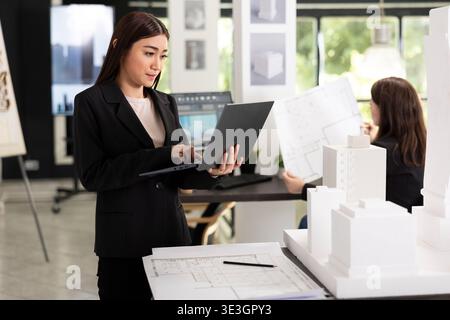 Professional designer reading digital blueprints and checking details on physical architectural model. Focused architect evaluating project requirements and creating reports for client presentations. Stock Photo