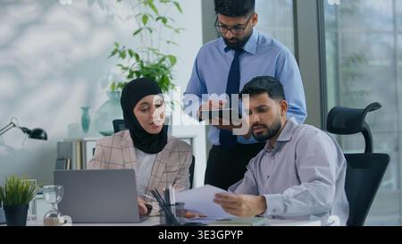 Team of business three Indian Arabian Muslim professionals in office discussing project strategy reviewing documents analyzing financial report Stock Photo