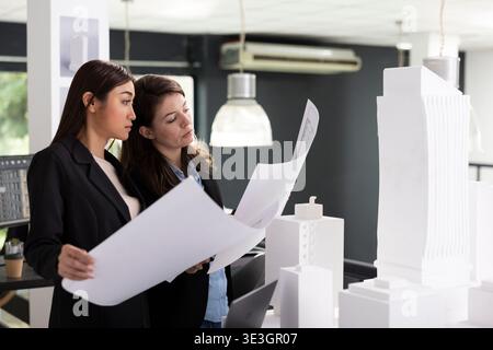Skilled architects study printed architectural blueprints, preparing for next construction site evaluation. Professional women crosschecking design variations for accurate project implementation. Stock Photo