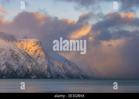 View of sunlight kissing snow-capped mountains as fog rolls in over the tranquil water, creating a serene yet dramatic landscape, Lyngen, Troms, Norwa Stock Photo