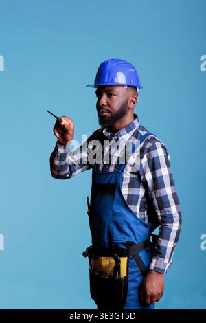 Portrait of construction professional from side holding screwdriver against blue background. Concentrated african american builder wearing tool belt and work uniform. Stock Photo