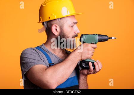 Male contractor holding electric drilling gun to screw nails and work on building project, standing over studio background. Handyman builder using cordless power drill tool, carpenter. Stock Photo