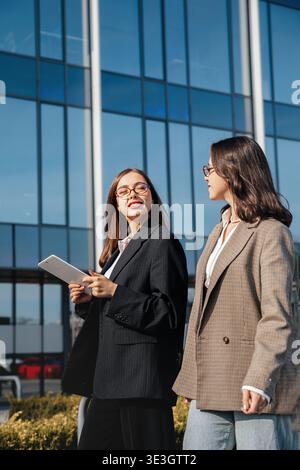 Two women walk outside a large office building while discussing work. They hold tablets and wear professional outfits. The sun shines brightly on them Stock Photo