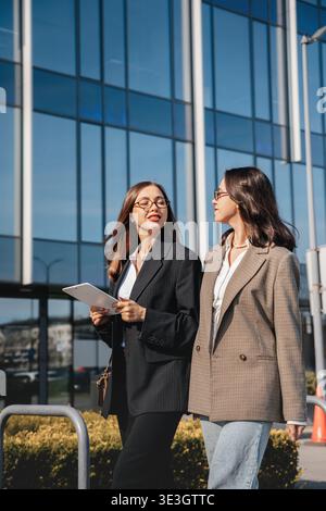Two women engage in conversation as they walk past a large office building. One holds a tablet while they discuss work and share ideas in the city. Stock Photo