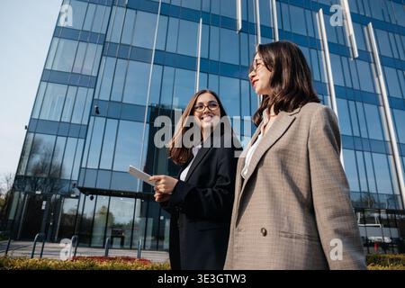 Two women in business attire are walking together outside a modern office building. They are engaged in conversation and appear focused on their work Stock Photo