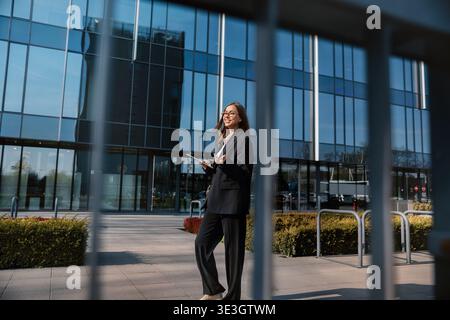 A woman in business attire stands outside a glass office building. She looks confident and engaged while using a tablet. The setting reflects a vibran Stock Photo