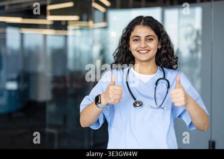 Young smiling woman medical worker wearing scrubs and a stethoscope, standing indoors, giving a confident thumbs up gesture, representing health success and care Stock Photo