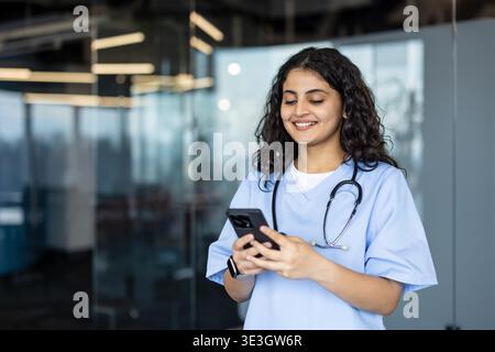 Young female nurse in scrubs smiles while checking her smartphone, managing telehealth communication and digital tasks in a modern clinic environment with confident professionalism Stock Photo