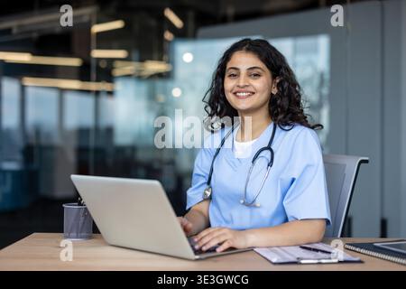 Young indian female doctor in scrubs with stethoscope sits at a desk in a modern clinic, smiling as she uses a laptop for telemedicine consultation and patient care Stock Photo