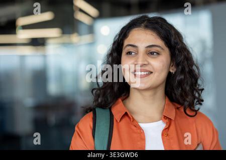 Young indian woman with curly hair wearing an orange shirt and backpack, smiling and looking away in a modern office or university setting, confident and hopeful about the future Stock Photo