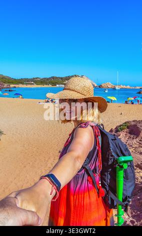 Woman in straw hat and summer dress smiling happily holding hands with a man guiding him towards the red sand beach of Cala Pregonda, Menorca, Spain. Stock Photo