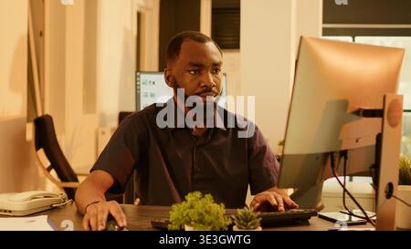 African american man working on research project with computer, analyzing data report to create presentation. Brainstorming ideas to plan management paperwork in office during sunset. Stock Photo