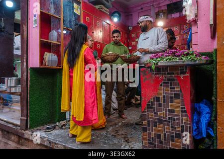 Customers in traditional clothing purchase floral offerings and snacks at a small, pink-walled shop in the Varanasi old town. Stock Photo
