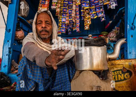 A street vendor in Varanasi sits inside his small blue wooden stall while preparing tea in a large metal kettle. Stock Photo