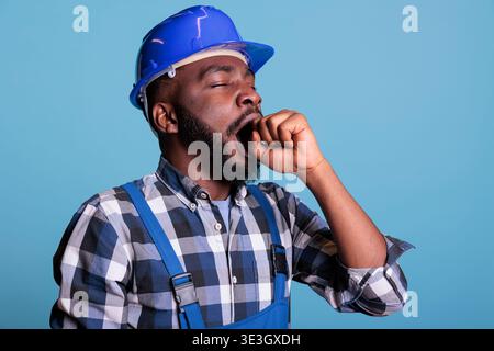 Exhausted african american man in coveralls and protective helmet isolated on blue background. Tired bearded construction worker tired with sleepiness after long working day. Stock Photo