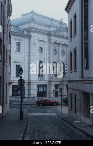 View of a vintage red car parked on cobblestones between old buildings under a misty sky, creating a timeless scene, Budapest, Hungary. Stock Photo