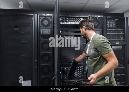 An IT technician uses a laptop to inspect and maintain server racks in a secure data center, performing network diagnostics, hardware checks and system monitoring in a professional environment. || Model released Stock Photo