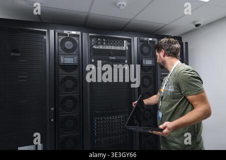 An IT technician inspects server racks in a modern data center while using a laptop to monitor equipment and perform maintenance. Focused technician works on network hardware in a secure server room. || Model released Stock Photo