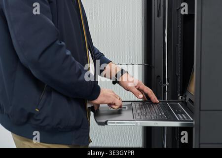 An IT technician types on a slide-out console laptop while configuring rack servers in a data center, checking network connections and performing maintenance on enterprise hardware and systems. Stock Photo