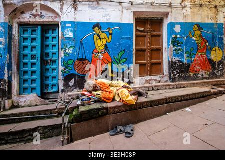 A man rests on a stone ledge in Varanasi before a wall decorated with blue and white paint and colourful murals depicting figures carrying loads. Stock Photo