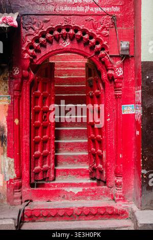 In Varanasi, a bright red carved stone archway featuring Hindu symbols opens onto a set of matching red stairs leading up into a building. Stock Photo