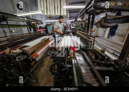 A worker operates a large mechanical power loom inside a textile workshop in Varanasi, where white fabric is being woven on industrial machinery. Stock Photo