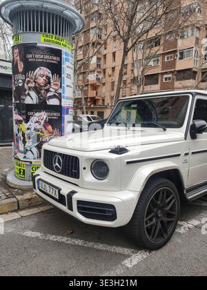 Close-up front three-quarter view of a white Mercedes-Benz G-Class G63 AMG parked on a city street in Barcelona next to a cylindrical advertising pill Stock Photo