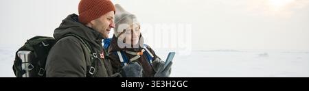 Header of middle aged Caucasian man and middle aged Caucasian woman standing outdoors in snowy landscape using smartphone together, both wearing winter hats and backpacks, looking at screen Stock Photo