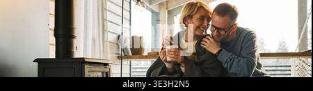 Header of middle aged Caucasian woman sitting beside middle aged Caucasian man holding mug, smiling and embracing near wood stove, both appearing relaxed and enjoying warm indoor moment together Stock Photo