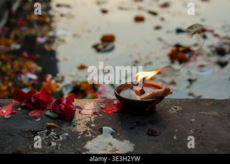 A traditional clay diya lamp burns on a stone surface in Varanasi next to red flower petals and offerings by the water. Stock Photo