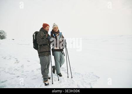 Adult Caucasian man and woman trekking through snowy landscape using hiking poles, wearing backpacks, smiling and looking at each other while walking outdoors Stock Photo