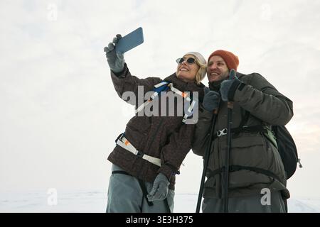 Caucasian woman and man standing outdoors in winter clothing smiling and taking selfie with smartphone, both wearing backpacks and gloves, snowy landscape in background Stock Photo