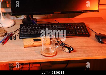 A close up view of a wooden desk surface featuring a keyboard, mouse, eyeglasses, a glass mug on a coaster, and a penknife, illuminated by warm ambien Stock Photo